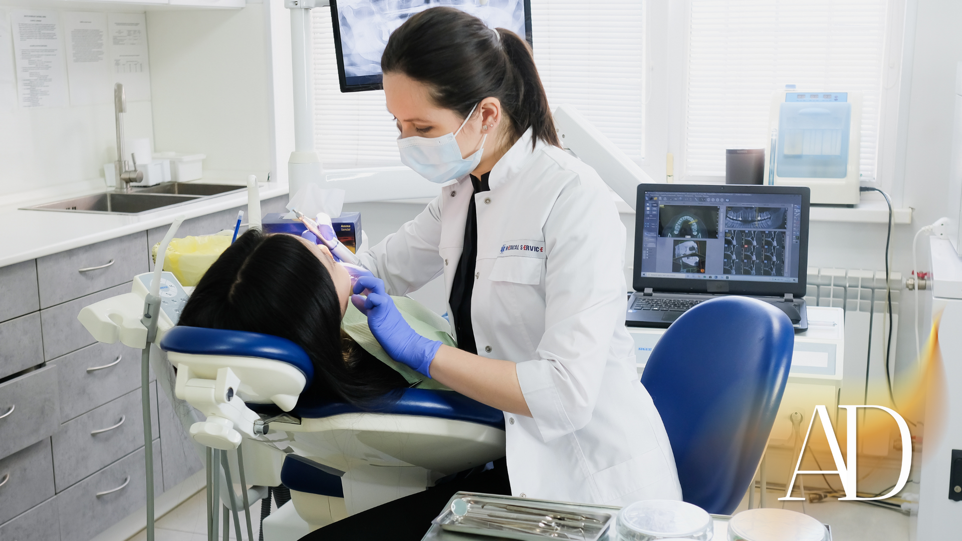 Dentist wearing a mask examines a patient in a dental chair beside a monitor and medical equipment.