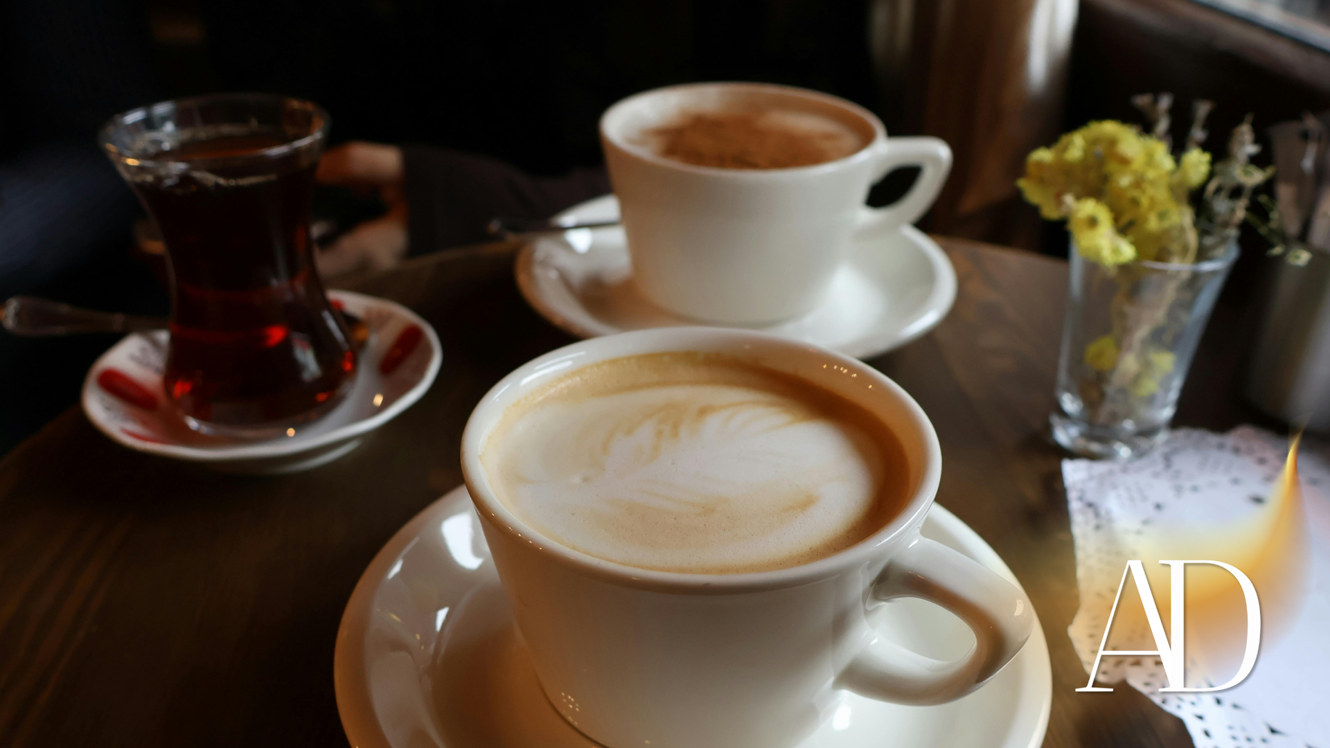 Coffee cups and a glass of tea on a table beside a small vase of yellow flowers.
