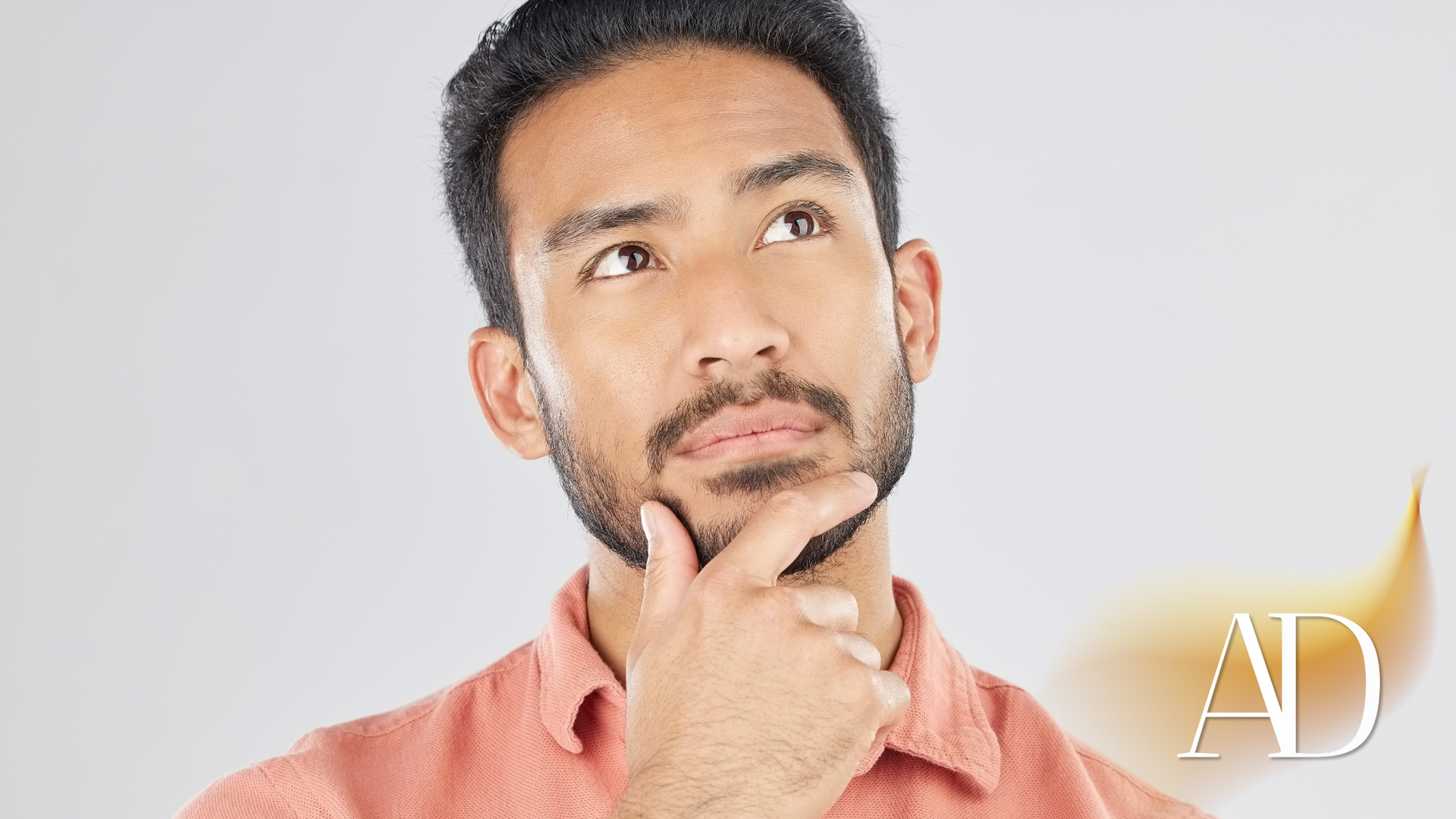 Man in a salmon shirt looking up thoughtfully with hand on chin against a white background