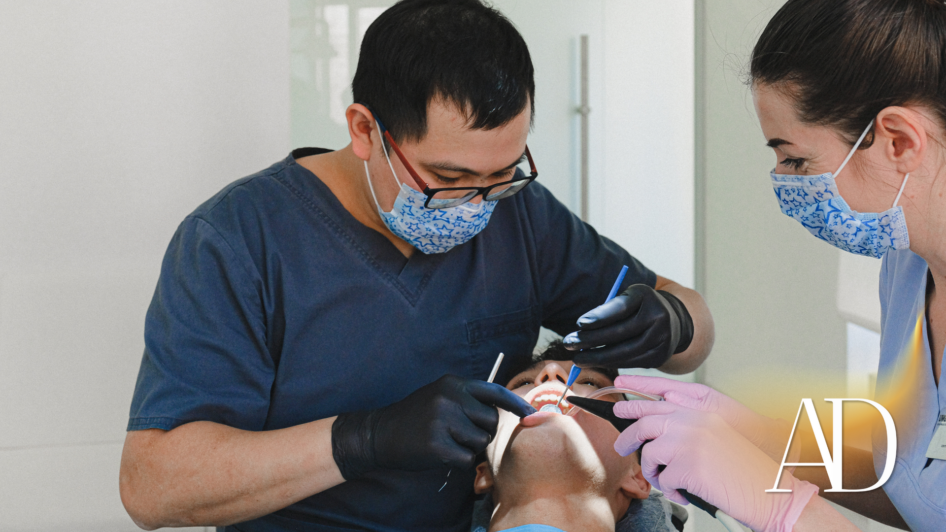 Dentist treating a patient while an assistant passes tools, all wearing masks and gloves in a clinic.