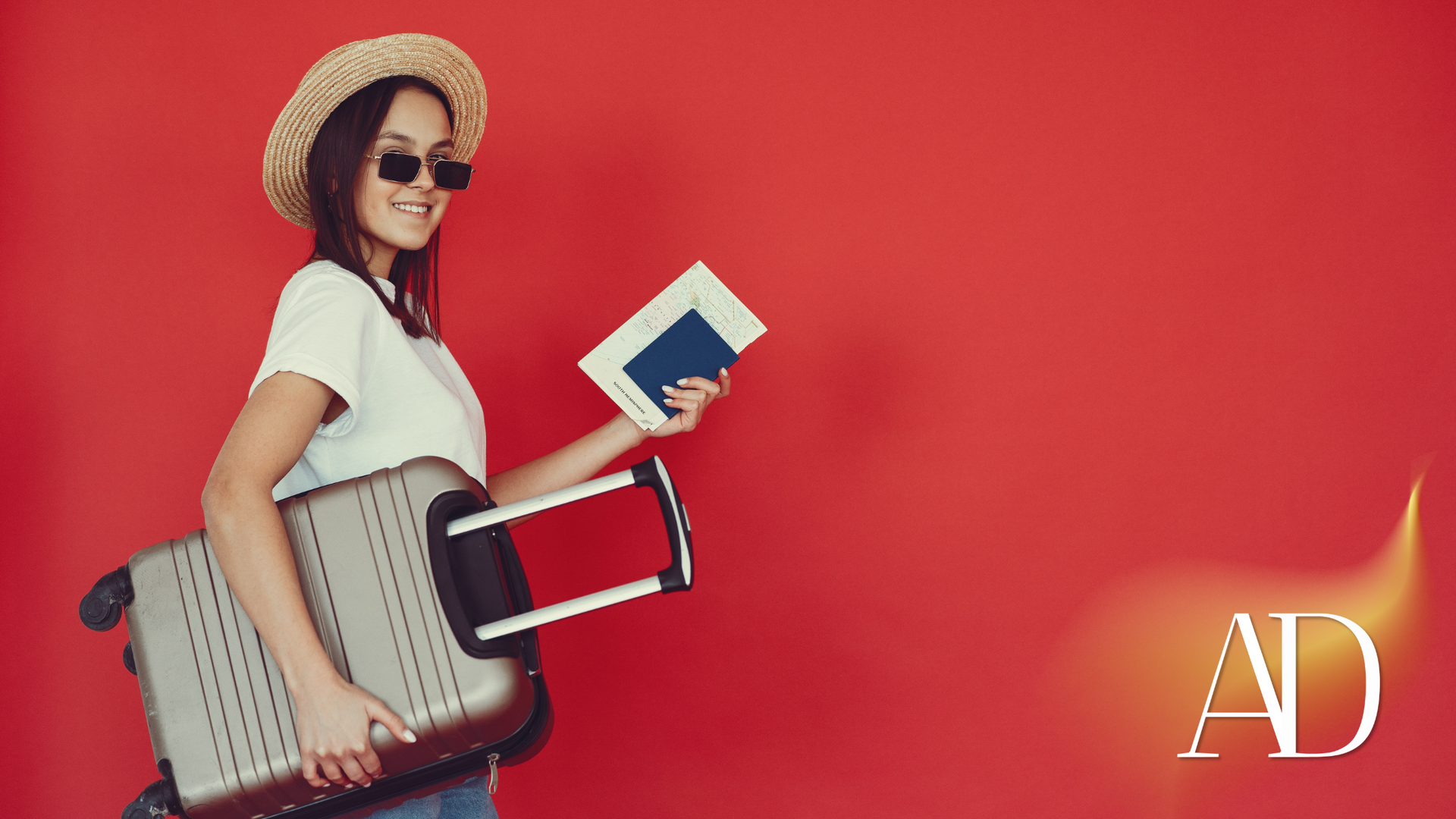 Smiling person in a straw hat and sunglasses holding a book and luggage against a red background.