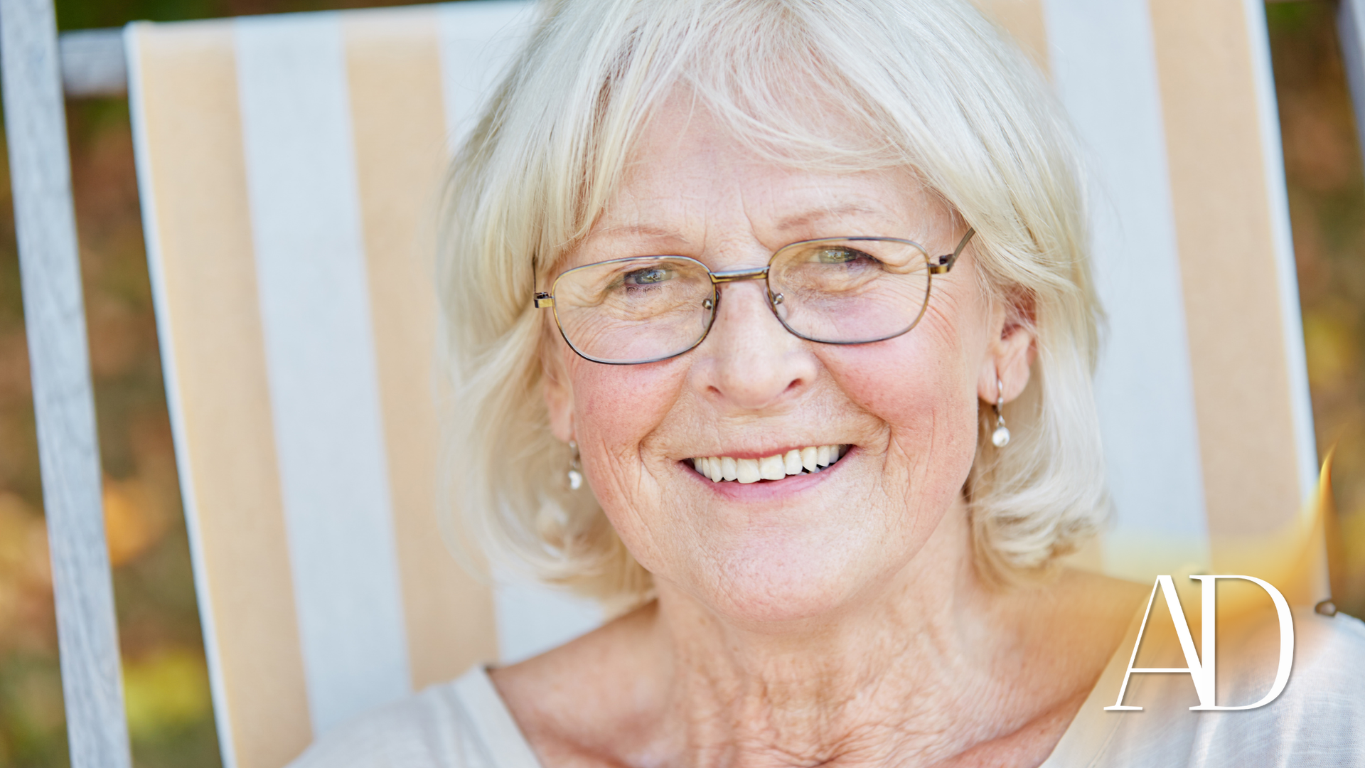 Smiling woman with white hair and glasses, seated against a striped background.