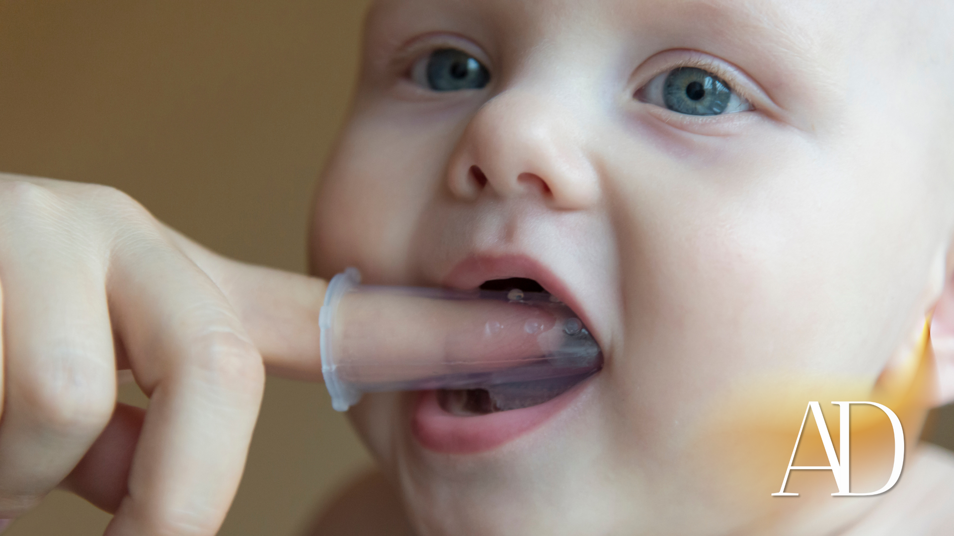 Close-up of a baby biting a transparent finger toothbrush, with a small AD logo in the corner.