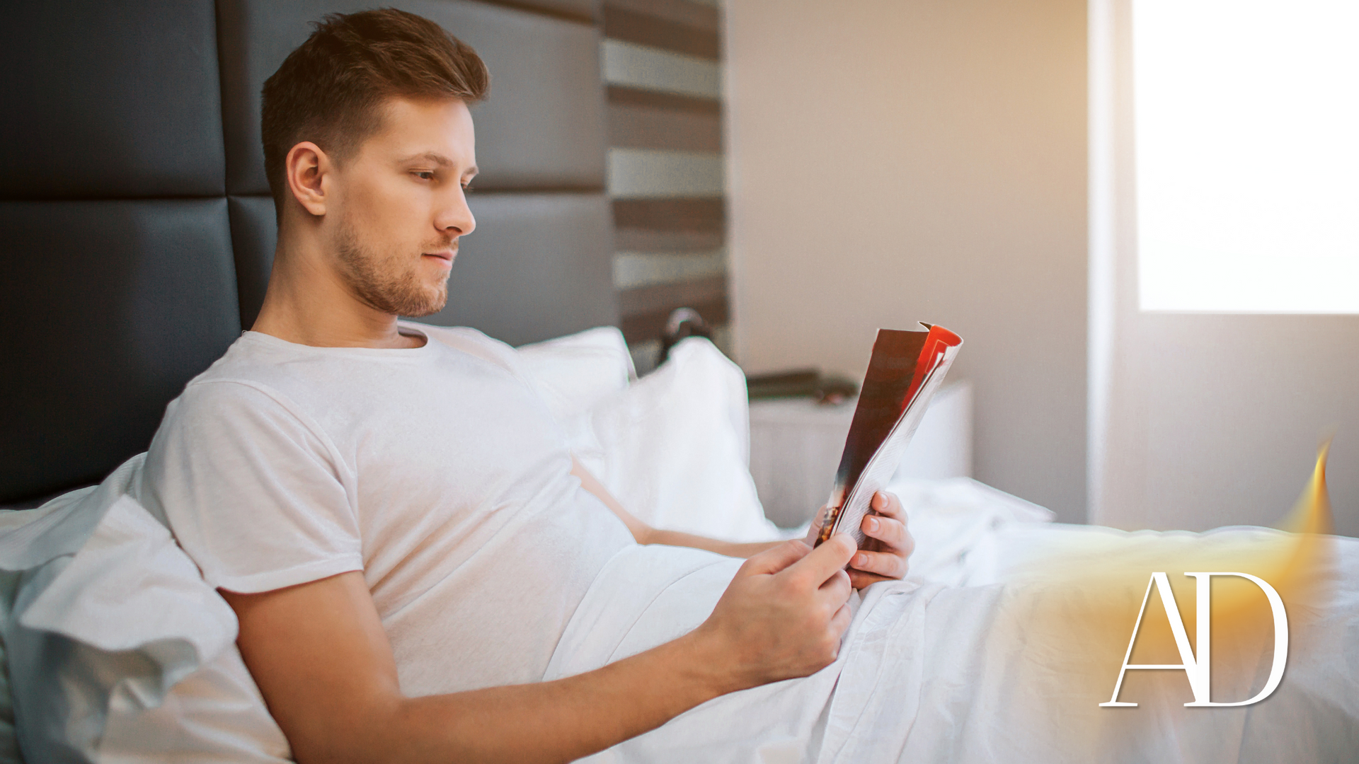 Person reclining in bed reading a magazine by a bright window