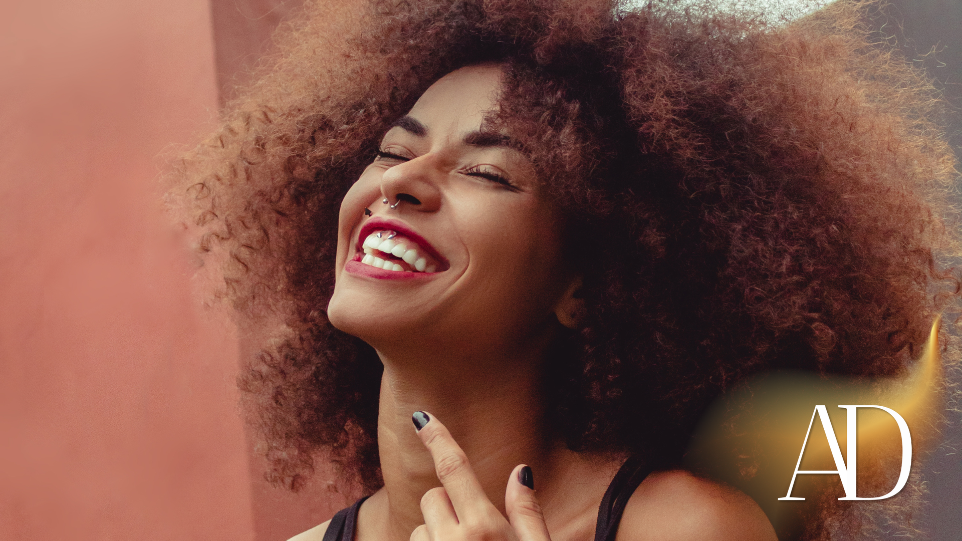 Smiling woman with curly hair against a warm pink background, laughing with head tilted back.