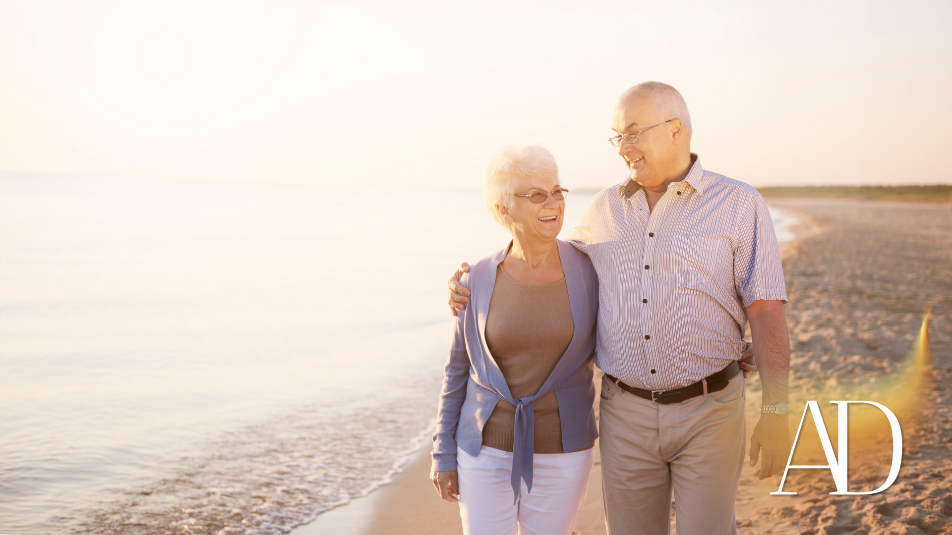 Two people walking hand in hand along a beach at sunset, with bright warm light.