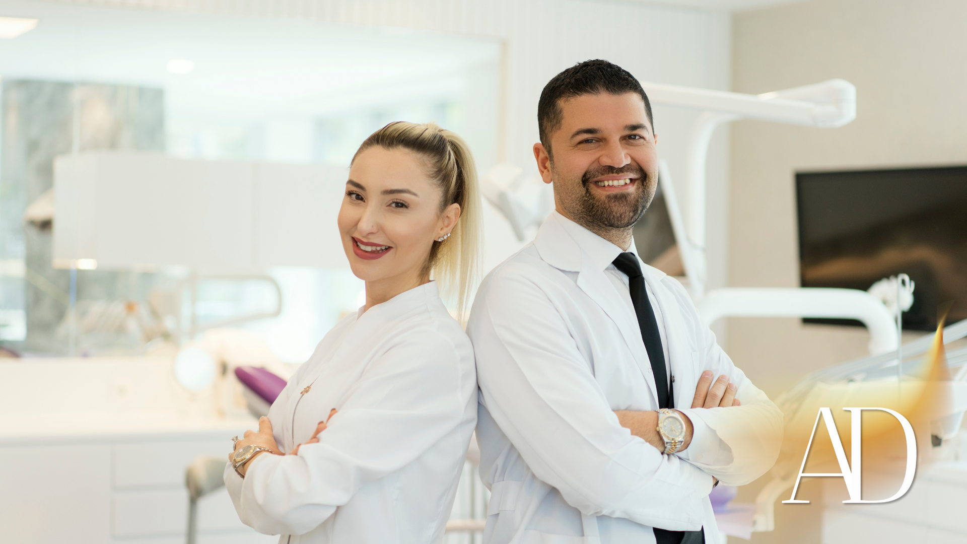 Two dental professionals in white coats stand back to back and smile in a bright clinic.