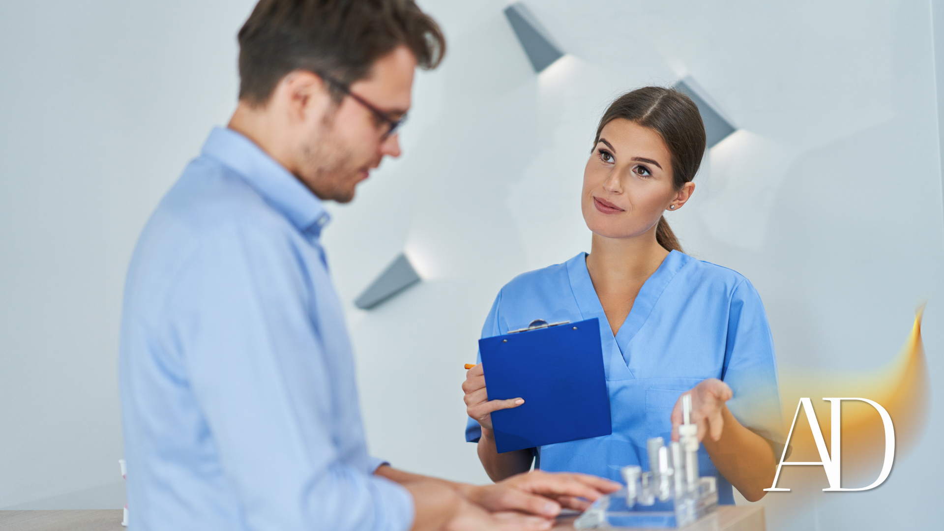 Two coworkers in a bright office, one holding a blue clipboard while talking to the other.