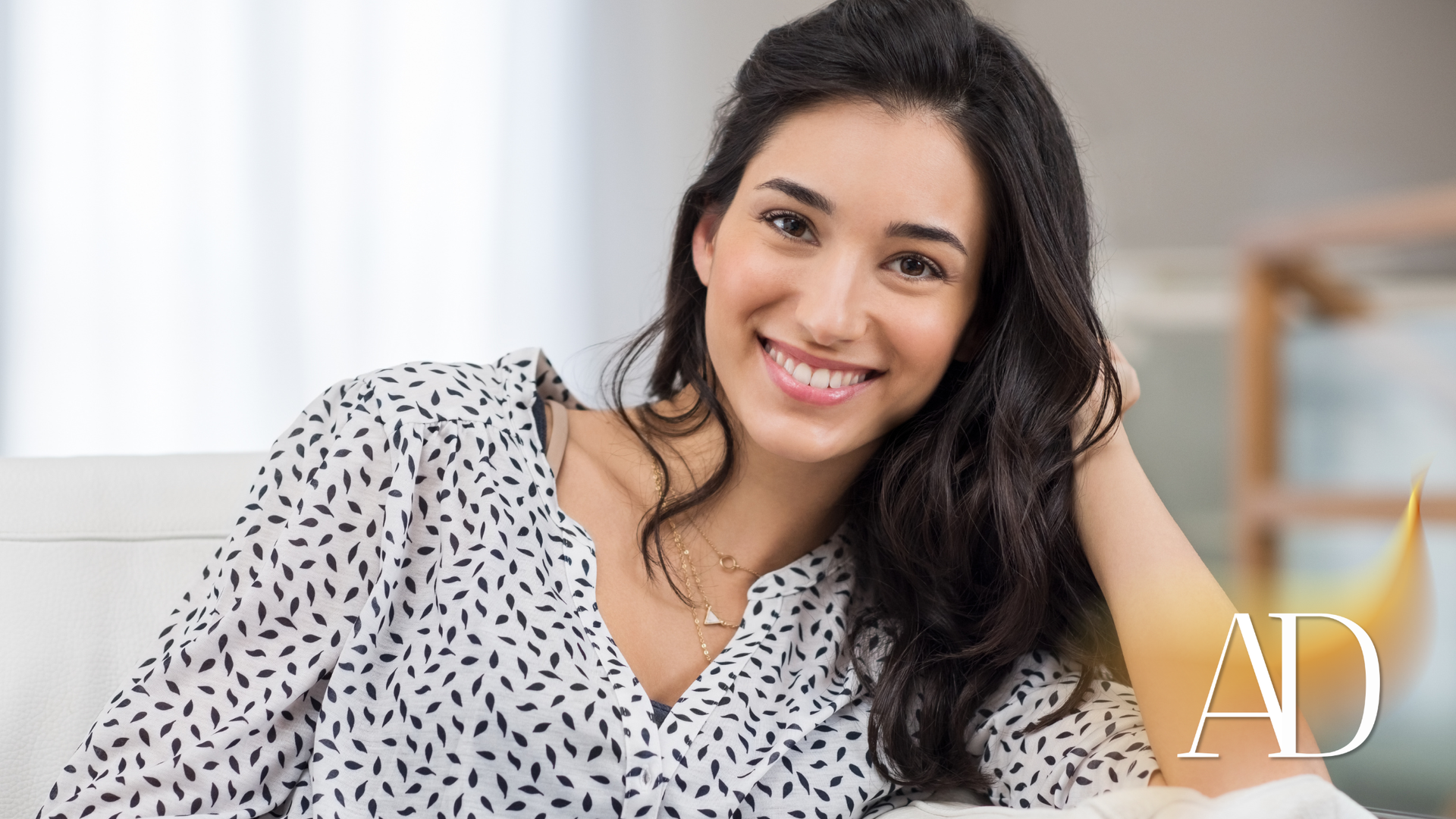 Smiling woman in a white patterned blouse reclines on a couch, resting her head on her hand