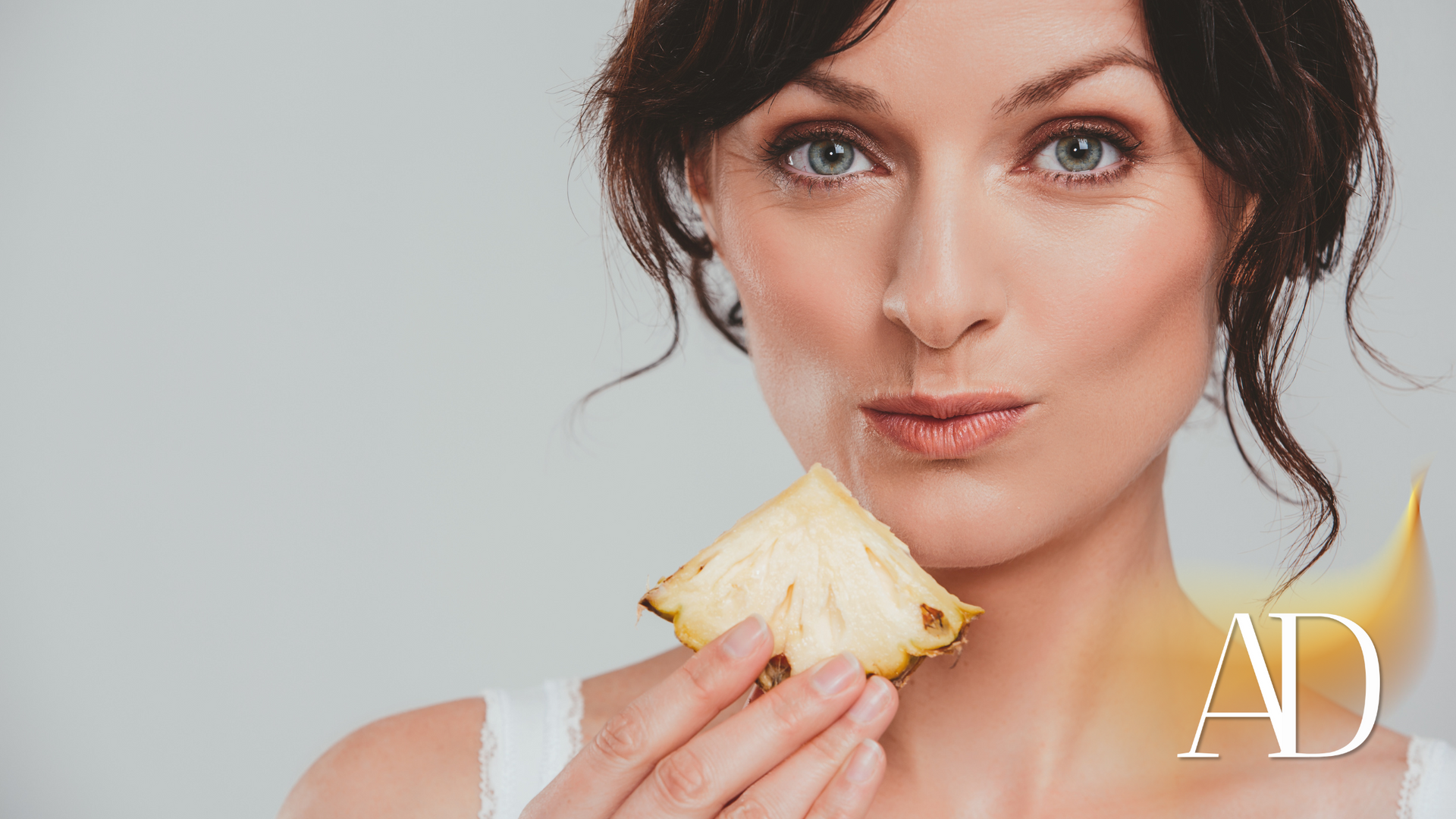 Woman with dark hair holding a sandwich and looking at the camera against a light background.