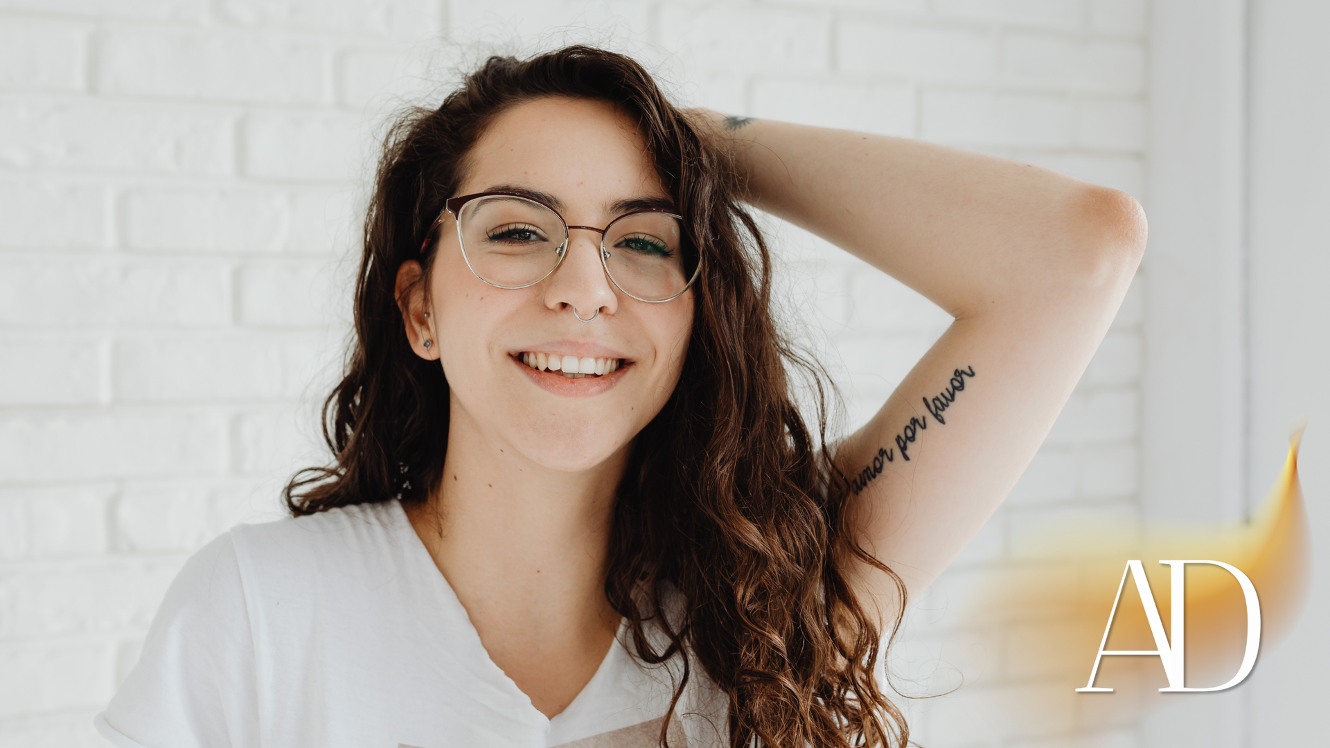 Smiling person with glasses and curly hair, reclining in a white shirt against a white brick wall