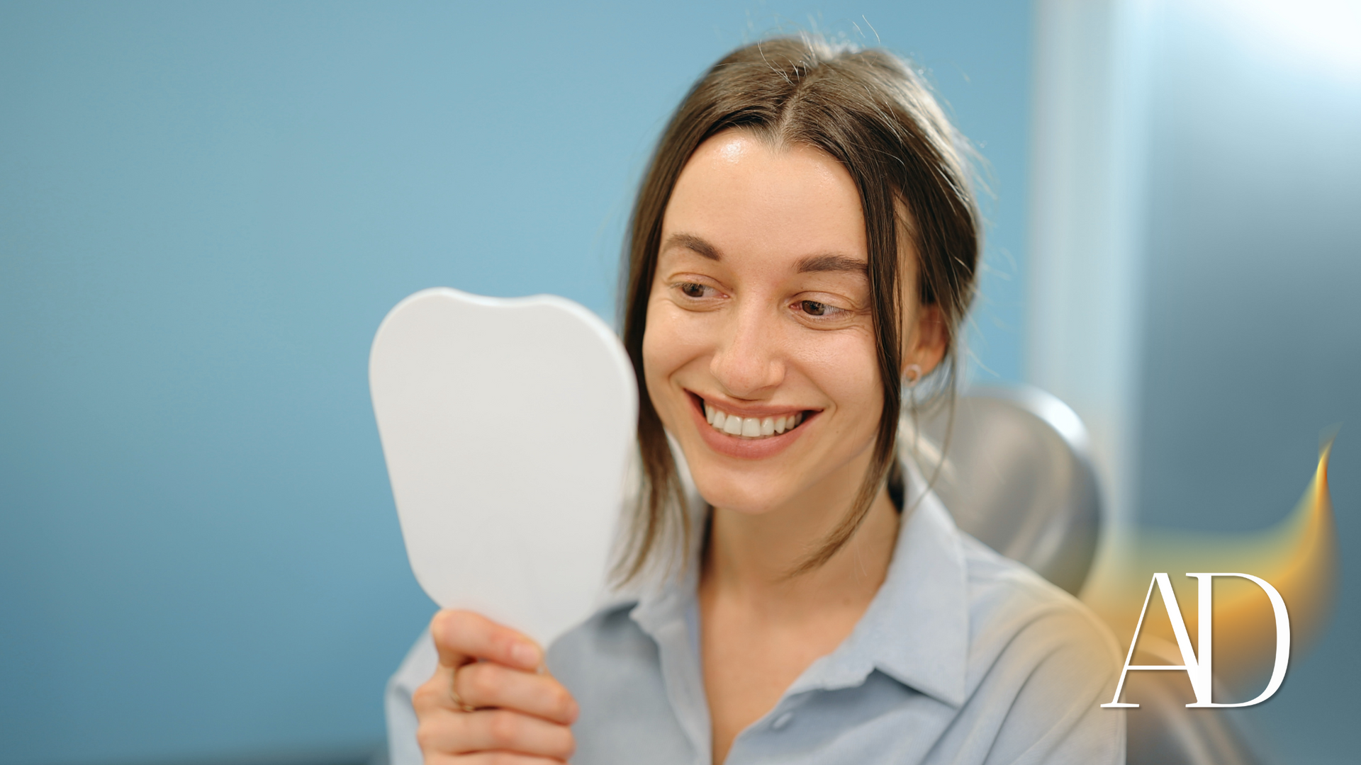 Woman smiling and holding a white dental shade guide in a bright clinic, with a blue background.