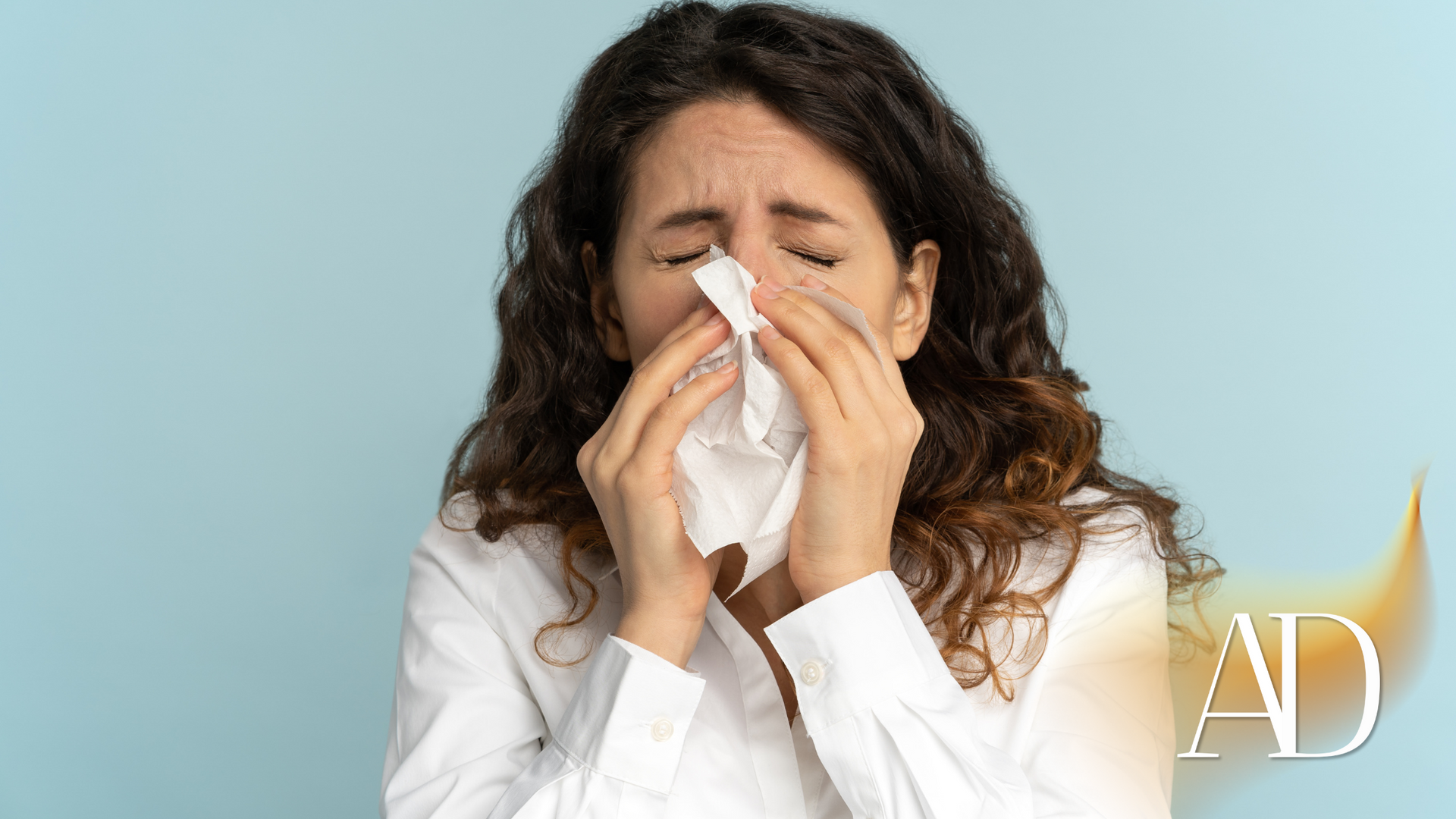 Woman in a white shirt blowing her nose into a tissue, eyes closed, against a light blue background.