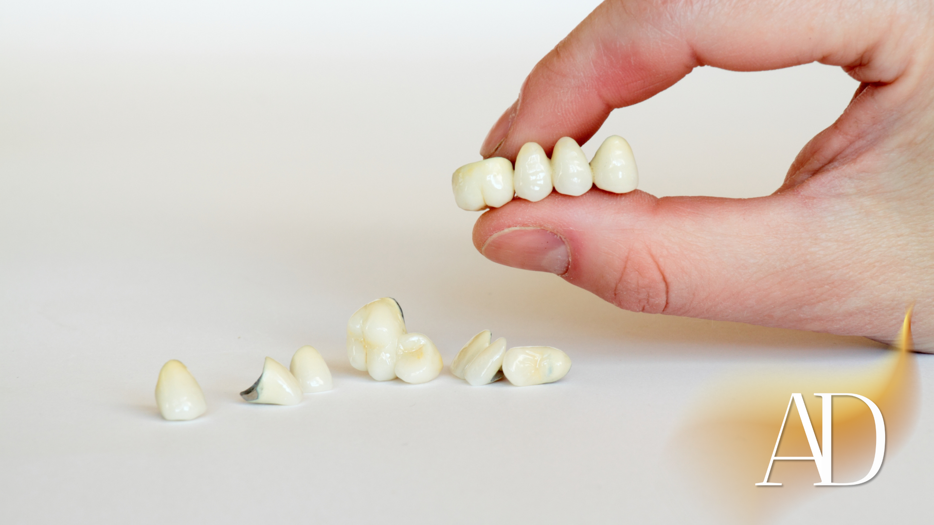 Hand holding a row of false teeth above several scattered dentures on a white surface