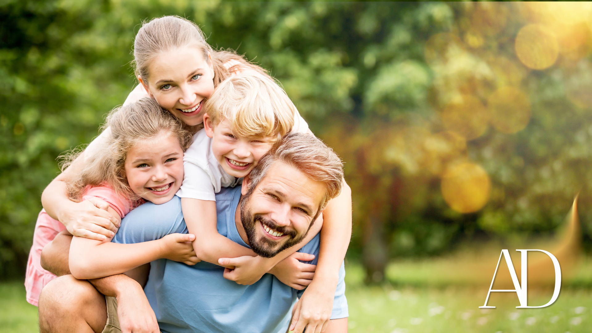 Smiling family of four outdoors in a park, with children hugging their parents in warm sunlight.