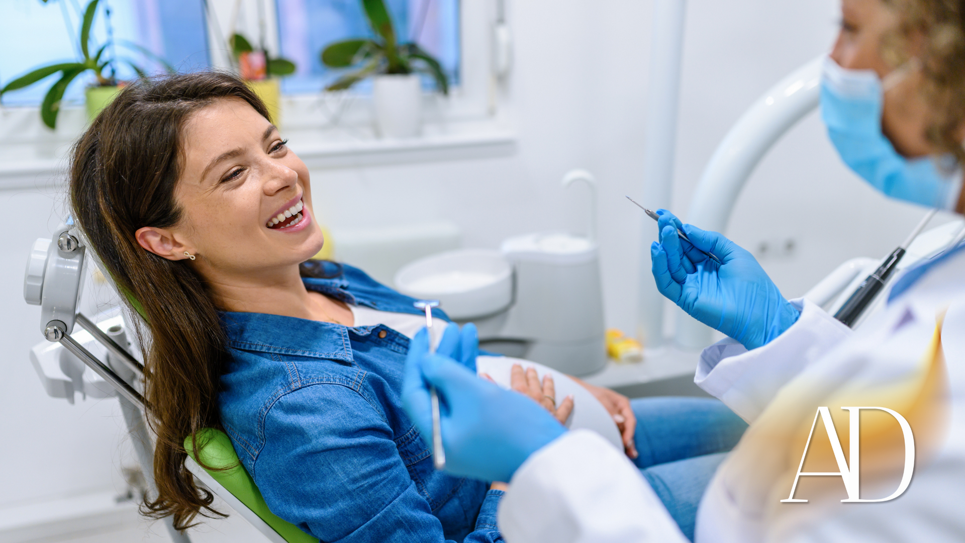 Smiling patient in blue top reclines in a dental chair as dentist prepares a procedure