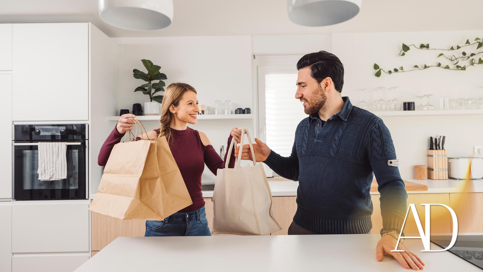 Two people in a bright kitchen clink drinks while holding grocery bags and smiling.