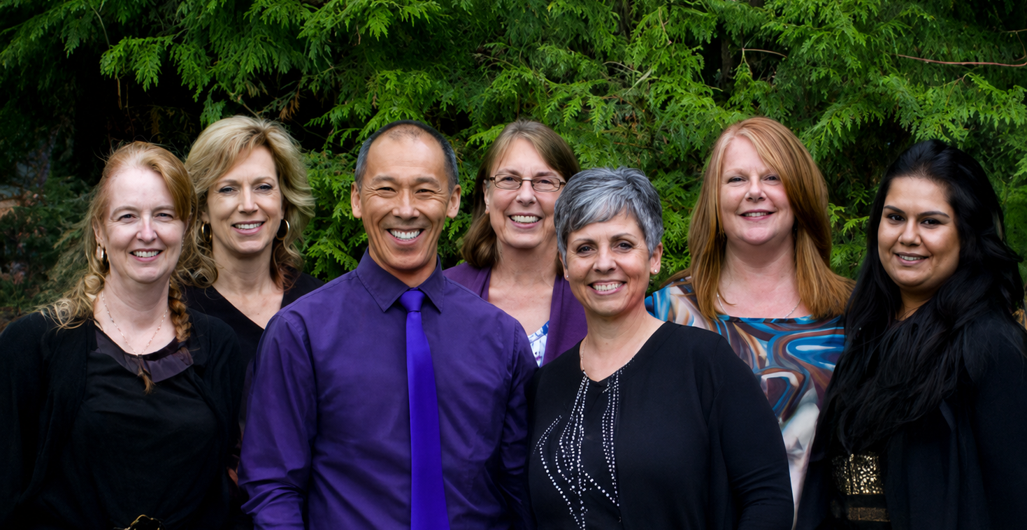Six smiling colleagues standing outdoors in front of green foliage, with one man in a purple shirt centered.