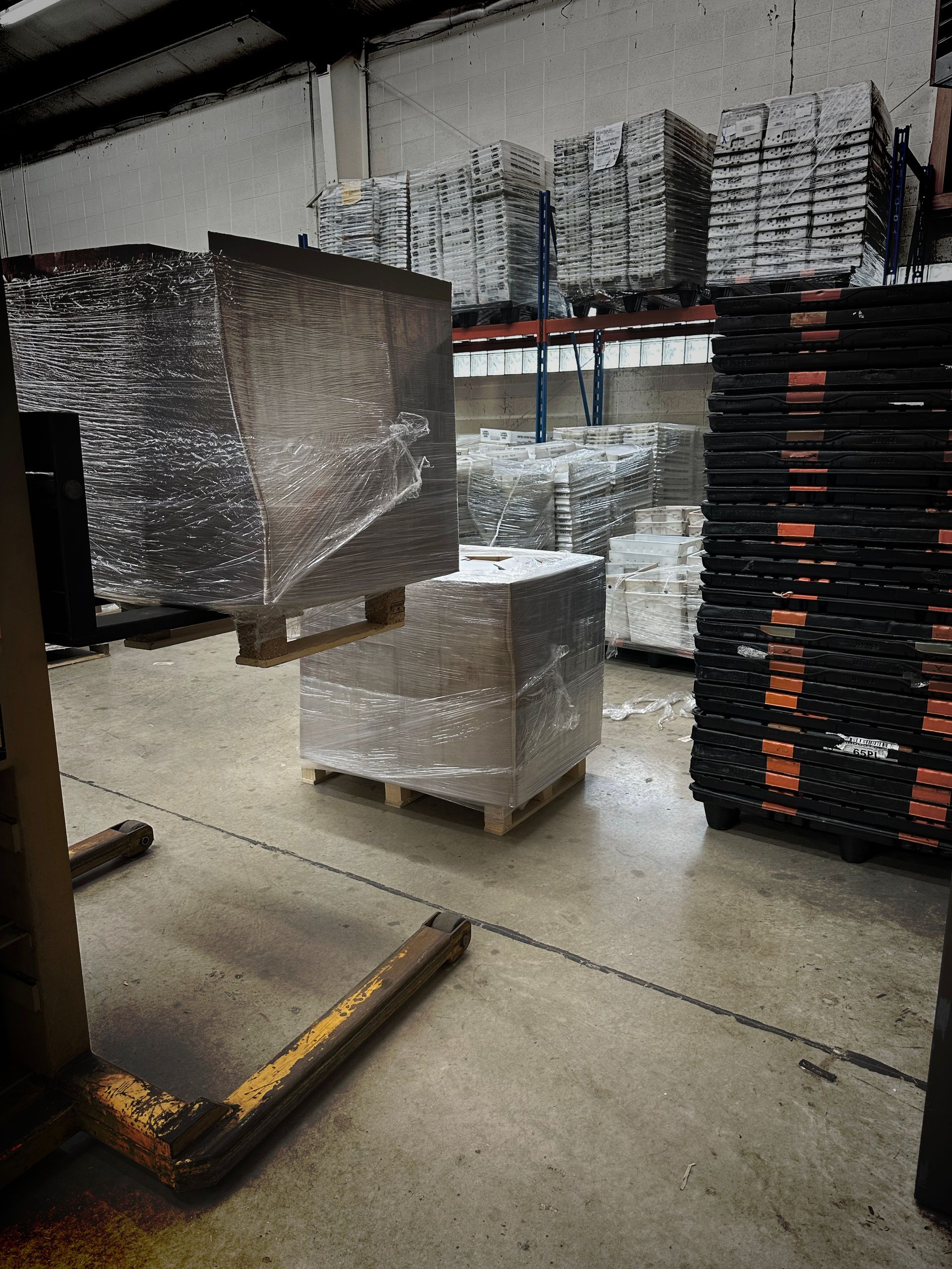 Pallets of shrink-wrapped boxes in a warehouse aisle, with stacked pallets in the background.
