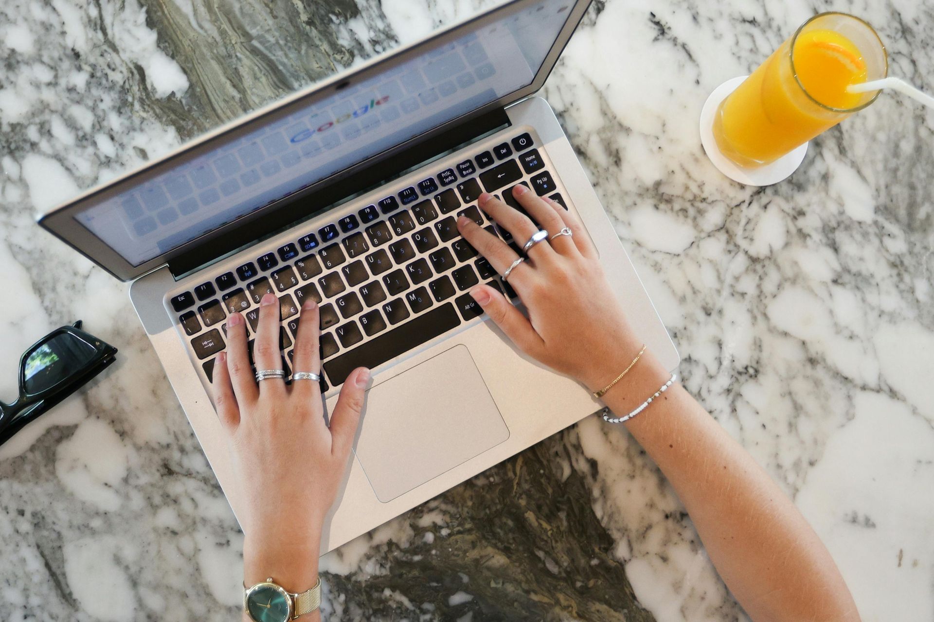 Hands typing on a laptop on a marble table beside a glass of orange juice.