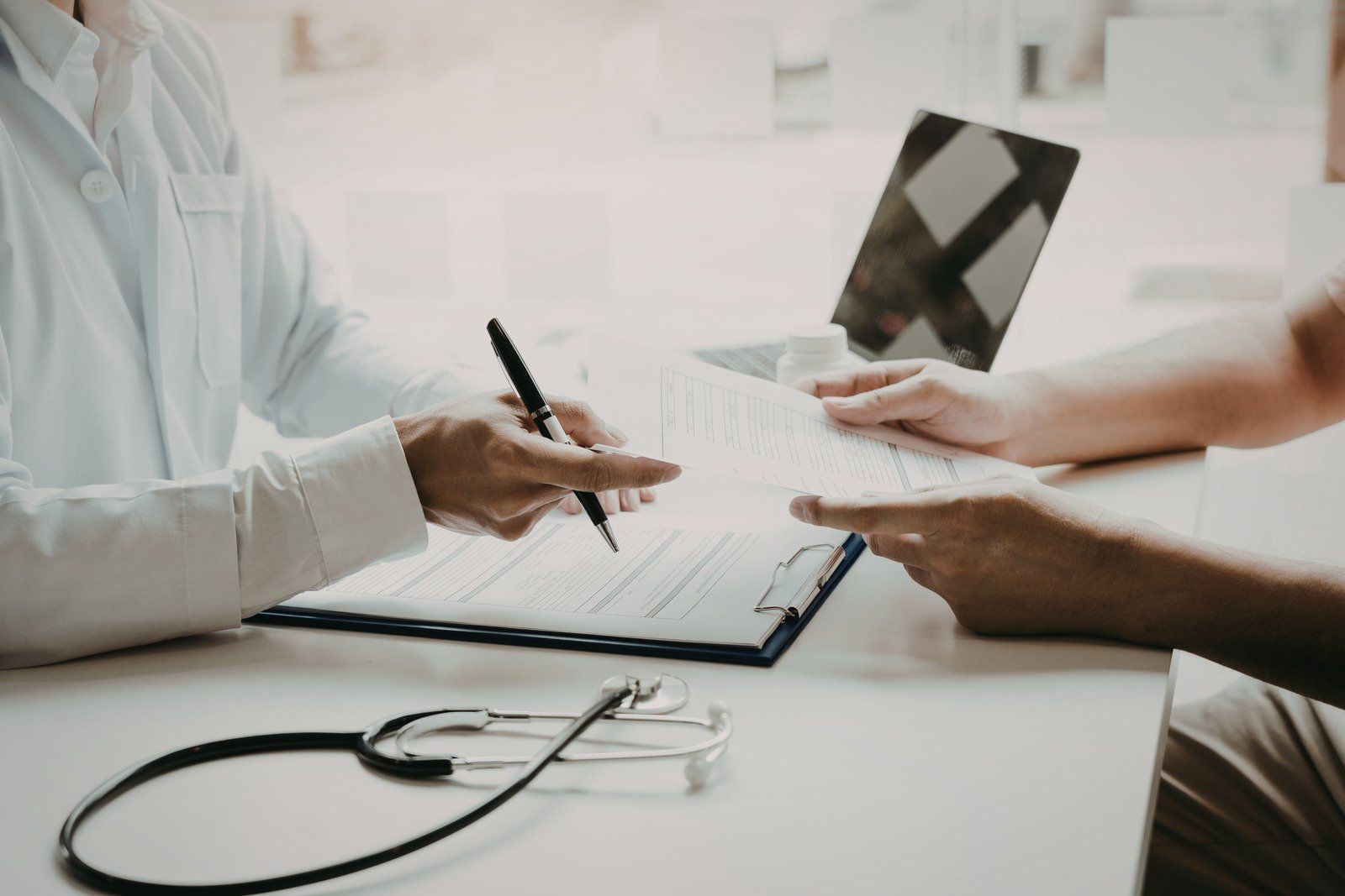 Doctor pointing at paperwork, discussing with patient; stethoscope rests on a white table.