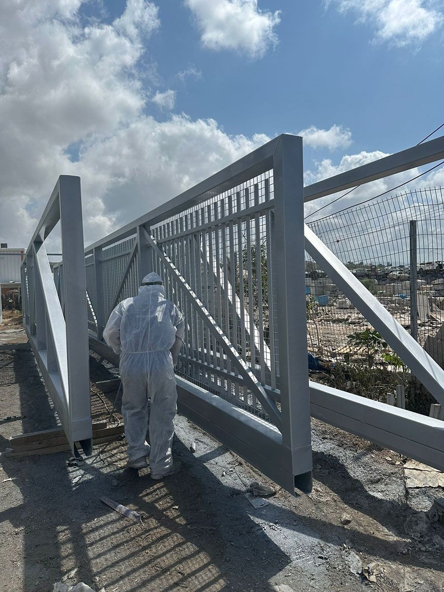 איש מקצוע בחליפת מגן מבצע צביעה תעשייתית קפדנית לשער, שלב הכרחי המבטיח עמידות לאורך שנים עבור שערים חשמליים קלים וכבדים.