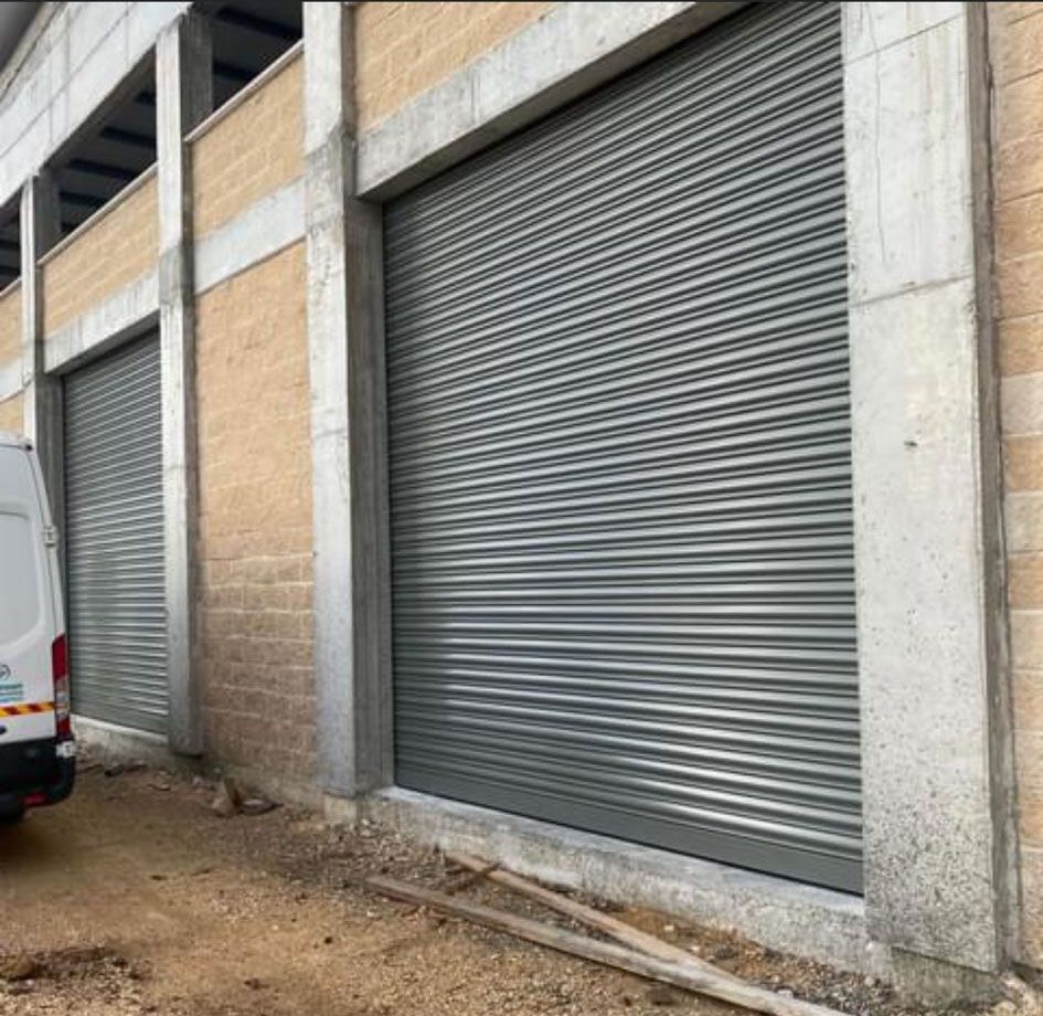 תמונה פנורמית המציגה סדרת תריסי גלילה מחוזקים על חזית מבנה תעשייתי, מתאים למרכזי לוגיסטיקה ומחסנים.