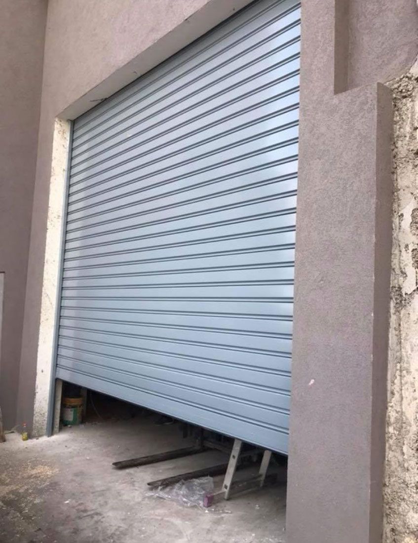תריסי גלילה שטוחים בצבע כסוף-אפור מותקנים בחזית מבנה בין קירות כהים.