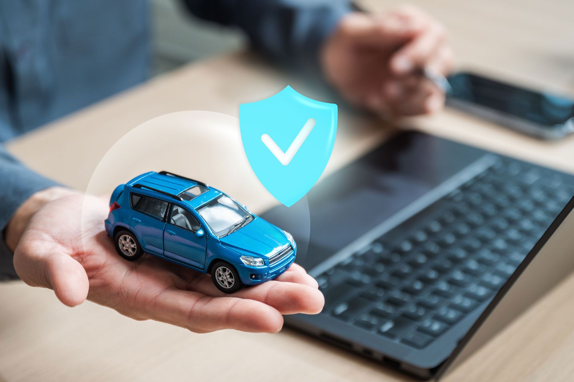 Blue toy car in a hand beside a laptop, with a shield icon indicating car insurance or security