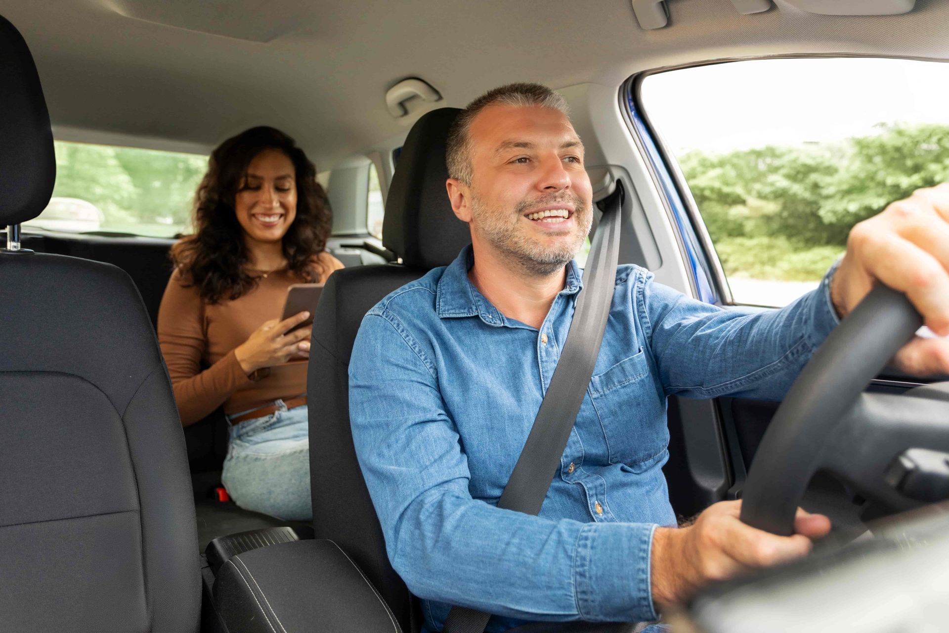 Man driving car, smiling, with passenger in the back using a phone.