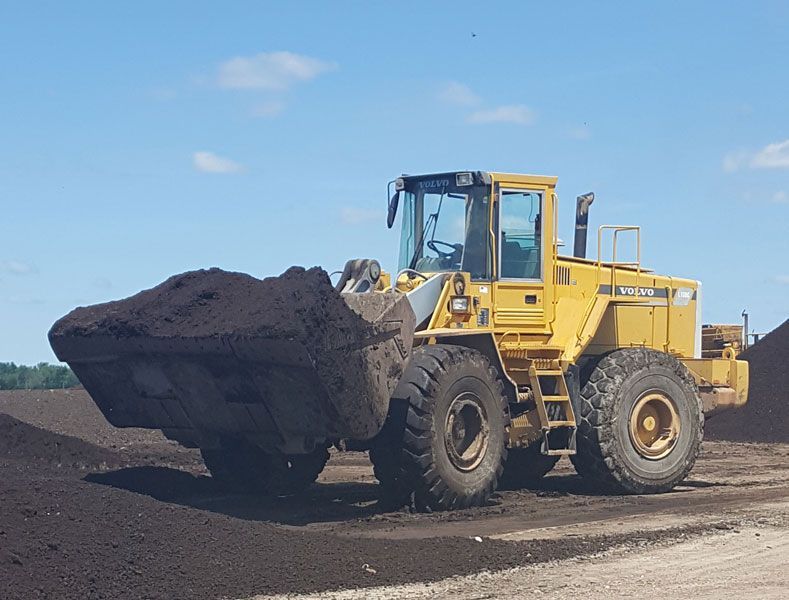 Yellow front loader hauling dark soil on a dirt site under a blue sky