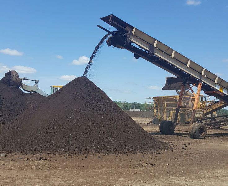 Conveyor pouring dark soil into a large pile at an outdoor industrial site
