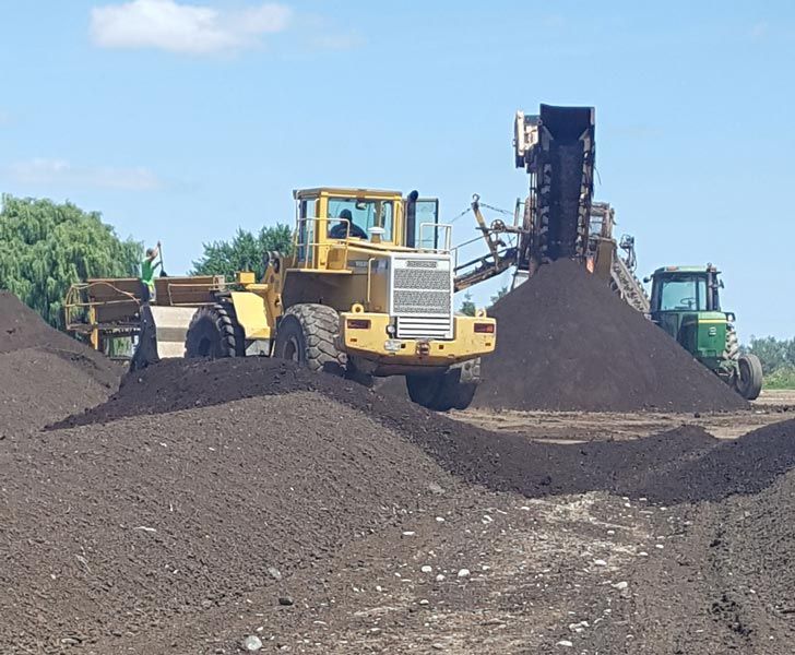 Yellow loader and green truck at a dark gravel quarry, with a conveyor piling material in the background.