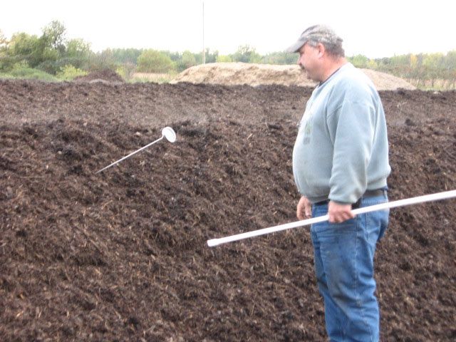 Man in a blue sweatshirt stands beside a large soil excavation with measuring rods.