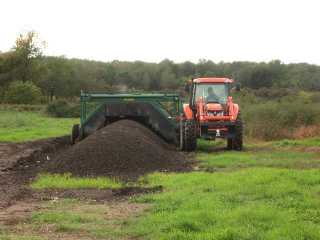 Orange tractor unloading dark soil onto a tarp in a grassy field under an overcast sky