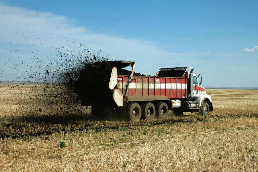White-and-red dump truck spreading black manure across a dry field under a blue sky