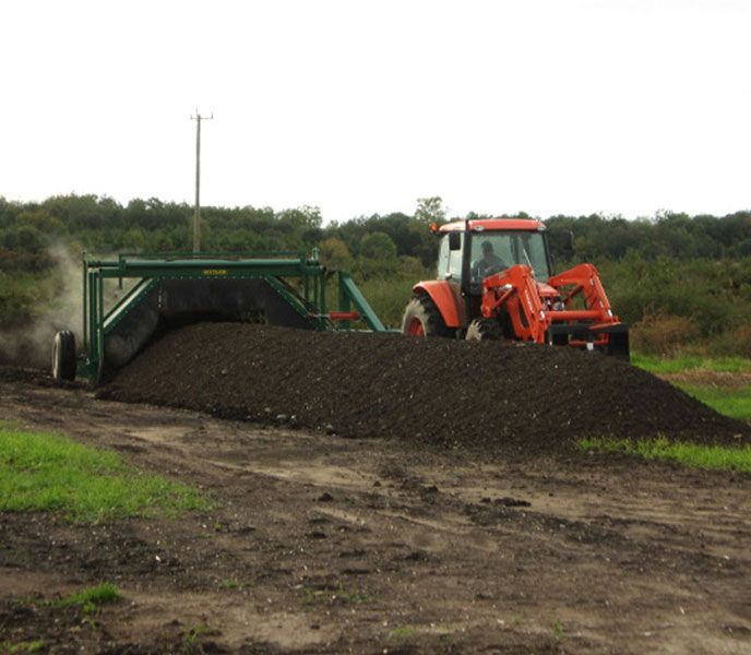 Orange tractor dumping a large pile of dark gravel beside a green machine on a rural dirt road