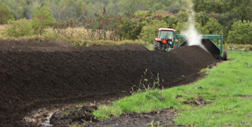 Tractor spreading dark compost along a field edge, with a plume of dust or steam nearby.