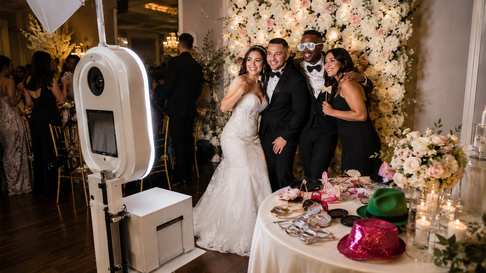Bride and groom posing with guests beside a photo booth at a floral-lit wedding reception