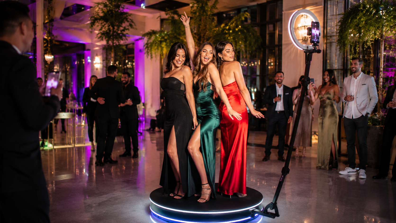 Three women in evening gowns pose on a lit platform at a formal event with photographers nearby