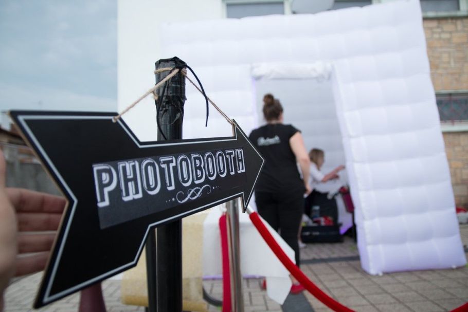 Black arrow sign reading “PHOTOBUD” beside a white inflatable photo booth at an outdoor event