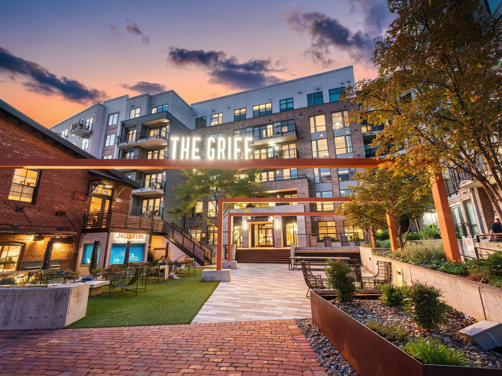 Courtyard entrance to The Griff Apartment at dusk, with lit windows, brick buildings, and trees.