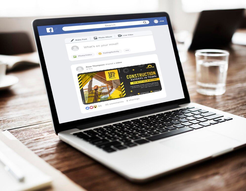 Silver laptop on a wooden table displaying a social media page, with a glass of water beside it