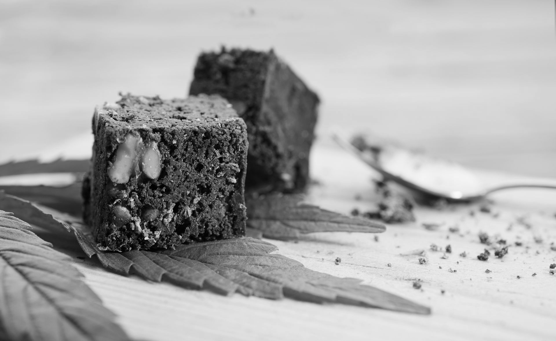 Two square brownies topped with nuts rest on a cannabis leaf on a wooden surface, with a spoon nearby.