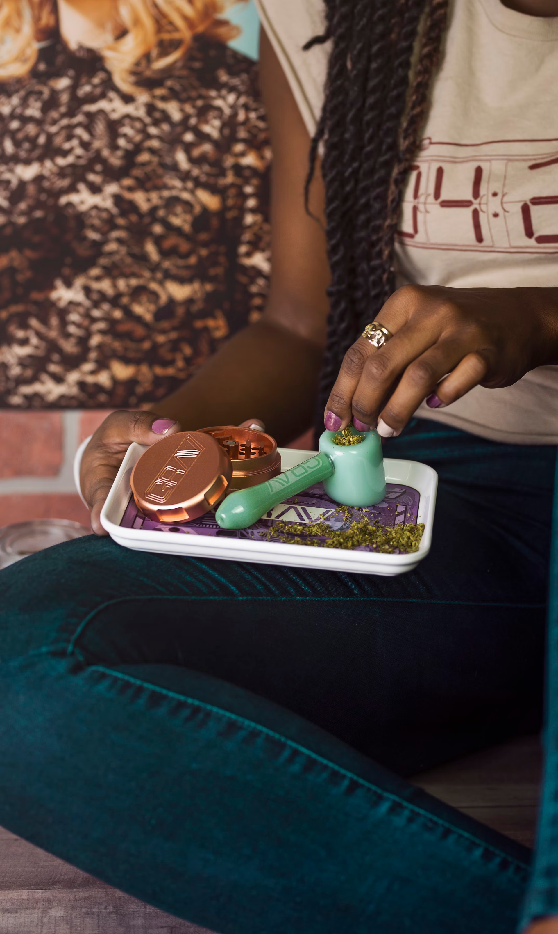 A person wearing a beige shirt and dark pants holds a white tray containing a metal grinder, a small pipe, and herbs.