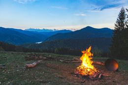Campfire burning brightly on a mountain top, with a dark blue mountain range in the background at dusk.