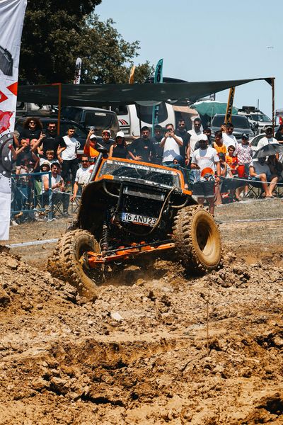 Orange off-road vehicle driving through muddy terrain during a competition, with spectators watching under a blue sky.