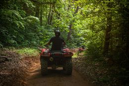 Person riding an ATV on a dirt path through a lush forest, with another ATV following.