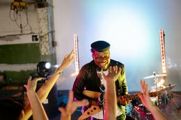 A musician plays guitar on stage, smiling with arms raised by an audience in a colorful lit venue.
