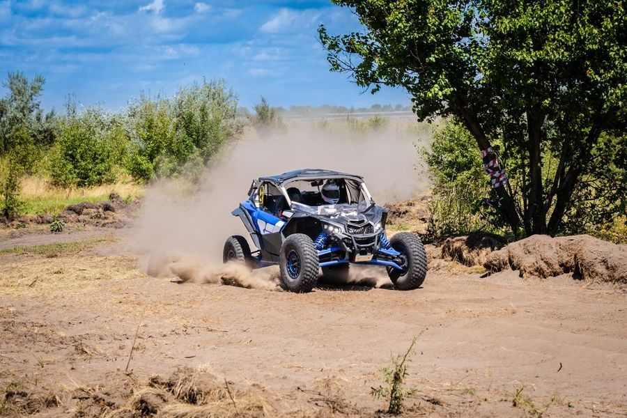 Blue and black off-road vehicle kicking up dust on a dirt track, trees in the background under a blue sky.